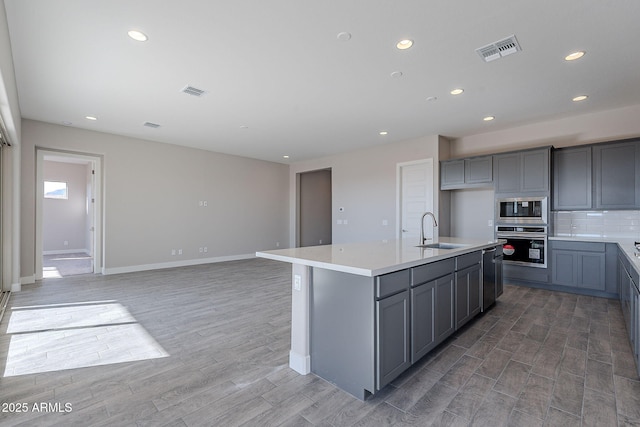 kitchen with a kitchen island with sink, sink, decorative backsplash, gray cabinets, and stainless steel appliances