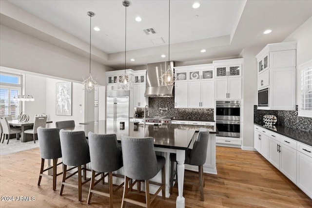 kitchen with built in appliances, a raised ceiling, a kitchen island with sink, and wall chimney exhaust hood