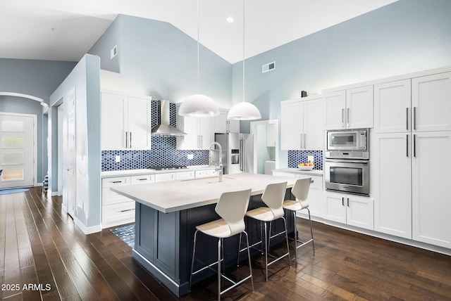 kitchen featuring wall chimney exhaust hood, white cabinetry, decorative light fixtures, appliances with stainless steel finishes, and an island with sink