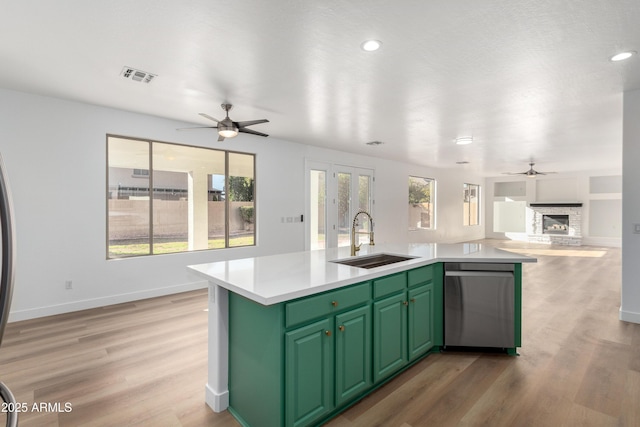 kitchen with sink, green cabinets, a stone fireplace, stainless steel dishwasher, and a kitchen island with sink