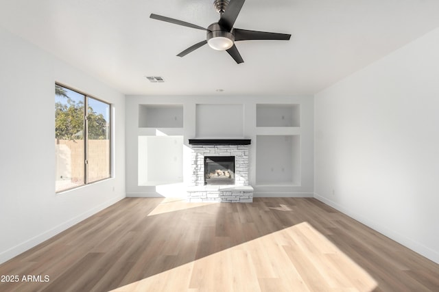 unfurnished living room with built in shelves, ceiling fan, a stone fireplace, and light wood-type flooring