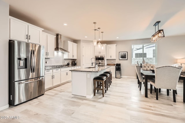 kitchen featuring dark stone counters, an island with sink, wall chimney exhaust hood, appliances with stainless steel finishes, and hanging light fixtures