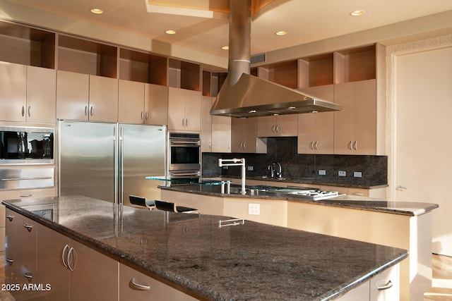 kitchen featuring wall chimney exhaust hood, tasteful backsplash, dark stone counters, a center island with sink, and appliances with stainless steel finishes