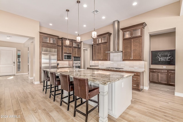 kitchen featuring decorative light fixtures, built in appliances, wall chimney exhaust hood, a large island, and light hardwood / wood-style floors