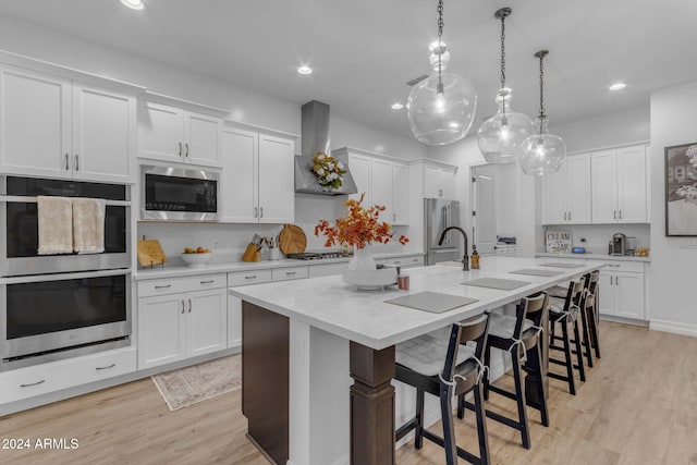 kitchen with a kitchen island with sink, light countertops, appliances with stainless steel finishes, white cabinetry, and wall chimney range hood