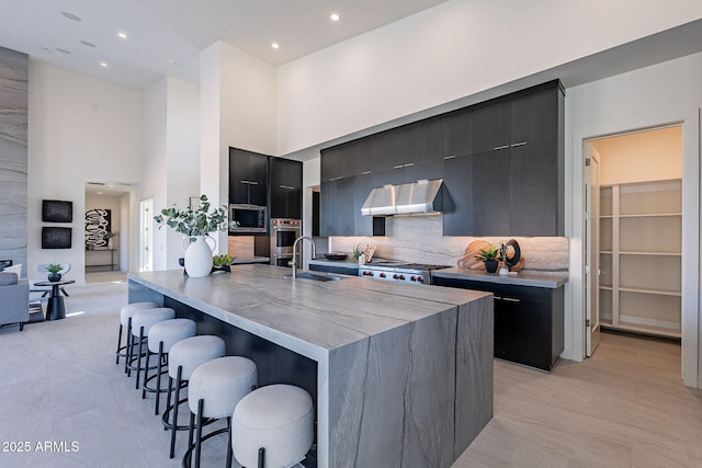 kitchen featuring a towering ceiling, stainless steel microwave, sink, a kitchen bar, and a kitchen island with sink