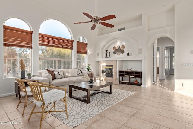 living room featuring a towering ceiling, a tiled fireplace, light tile patterned flooring, and ceiling fan