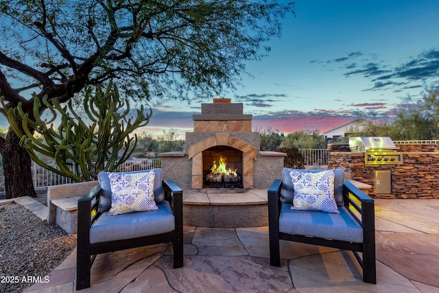 patio terrace at dusk featuring an outdoor kitchen, an outdoor stone fireplace, and area for grilling