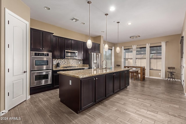 kitchen featuring pendant lighting, a center island with sink, light hardwood / wood-style flooring, light stone counters, and stainless steel appliances