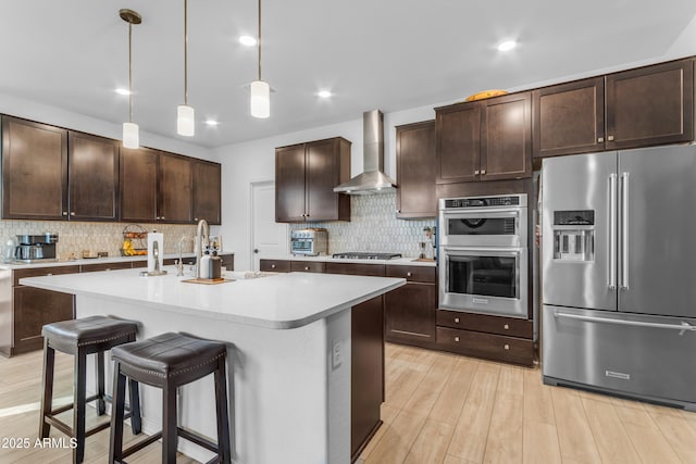 kitchen featuring dark brown cabinets, an island with sink, pendant lighting, stainless steel appliances, and wall chimney range hood