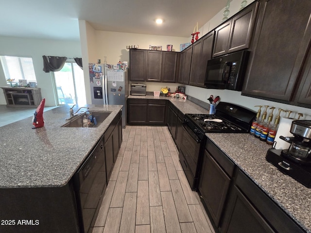 kitchen with sink, stone counters, a kitchen island with sink, dark brown cabinets, and black appliances