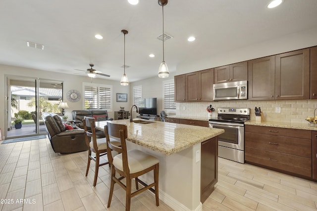 kitchen featuring a kitchen island with sink, hanging light fixtures, stainless steel appliances, light stone counters, and a kitchen bar