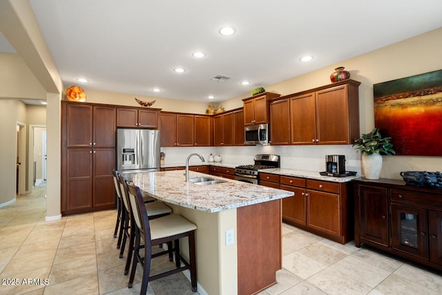 kitchen with a breakfast bar, an island with sink, a sink, light stone counters, and stainless steel appliances