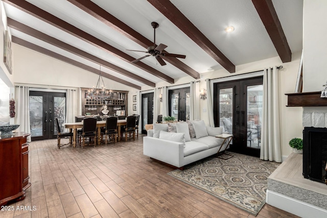 living area featuring lofted ceiling with beams, wood finished floors, french doors, a fireplace, and ceiling fan with notable chandelier