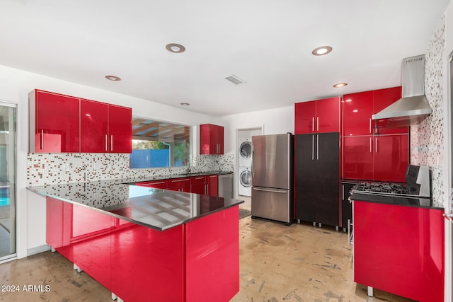 kitchen featuring decorative backsplash, kitchen peninsula, stainless steel appliances, wall chimney range hood, and stacked washer and dryer