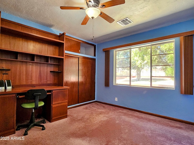 unfurnished office with a textured ceiling, visible vents, built in desk, a ceiling fan, and carpet floors
