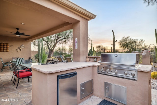 patio terrace at dusk with ceiling fan, an outdoor kitchen, a grill, and a bar