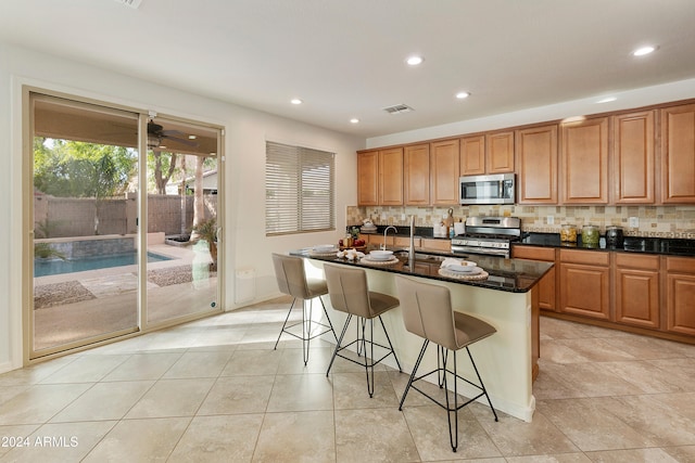 kitchen featuring backsplash, appliances with stainless steel finishes, an island with sink, a kitchen bar, and dark stone countertops