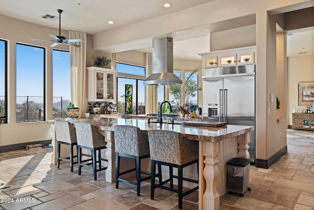 kitchen with a breakfast bar, a center island with sink, white cabinets, built in refrigerator, and island exhaust hood