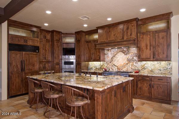 kitchen featuring a center island with sink, light stone counters, tasteful backsplash, a breakfast bar area, and stainless steel double oven