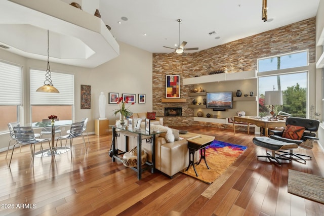 living room featuring a stone fireplace, a high ceiling, wood finished floors, and ceiling fan