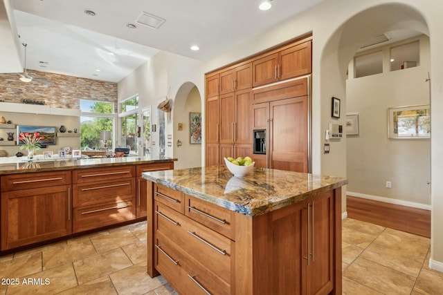 kitchen featuring stone countertops, a kitchen island, arched walkways, brown cabinetry, and paneled built in refrigerator