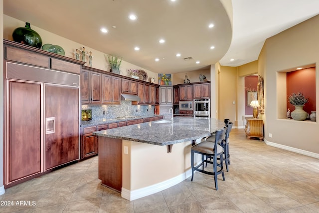 kitchen featuring sink, built in appliances, dark stone counters, a spacious island, and a kitchen bar