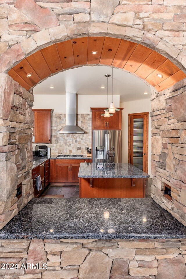 kitchen with dark stone countertops, backsplash, stainless steel appliances, hanging light fixtures, and wall chimney exhaust hood