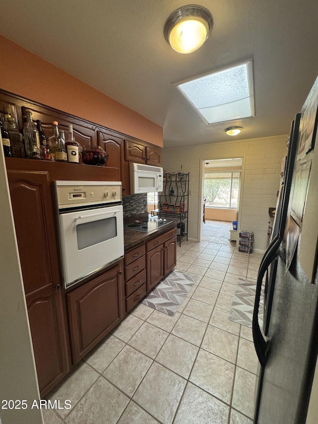 kitchen featuring light tile patterned floors, dark countertops, dark brown cabinets, vaulted ceiling with skylight, and white appliances