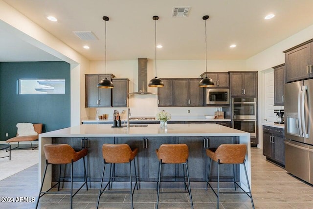 kitchen featuring wall chimney exhaust hood, light hardwood / wood-style floors, a center island with sink, decorative light fixtures, and appliances with stainless steel finishes