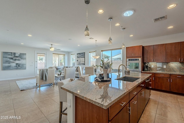 kitchen featuring backsplash, a kitchen island with sink, ceiling fan, sink, and hanging light fixtures