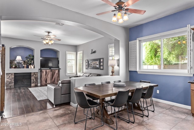tiled dining space featuring ceiling fan, a fireplace, arched walkways, and baseboards