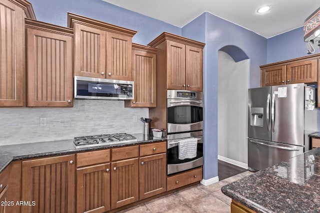 kitchen featuring stainless steel appliances, dark stone countertops, tasteful backsplash, arched walkways, and brown cabinetry