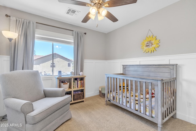 carpeted bedroom featuring a wainscoted wall, visible vents, a decorative wall, ceiling fan, and a crib