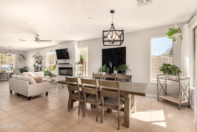 tiled dining room with ceiling fan with notable chandelier and a stone fireplace