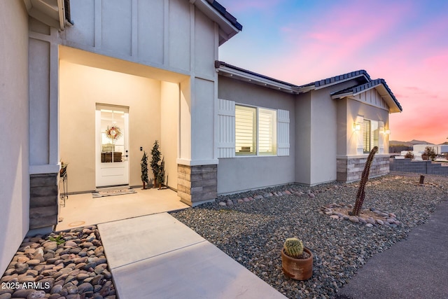 property exterior at dusk featuring stone siding, board and batten siding, a patio area, and stucco siding