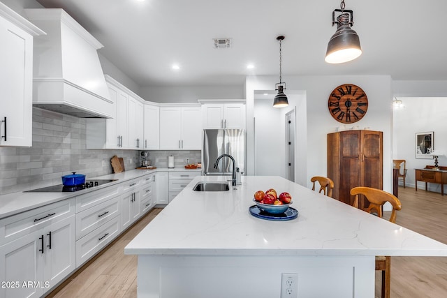 kitchen featuring hanging light fixtures, a kitchen island with sink, a sink, stainless steel fridge, and premium range hood