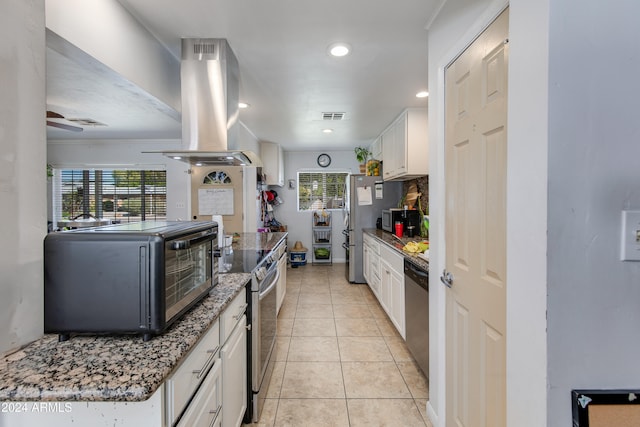 kitchen featuring white cabinets, light tile patterned floors, appliances with stainless steel finishes, island range hood, and dark stone countertops