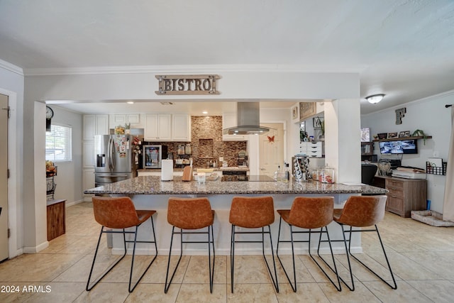 kitchen with wall chimney exhaust hood, dark stone countertops, ornamental molding, stainless steel fridge, and white cabinets