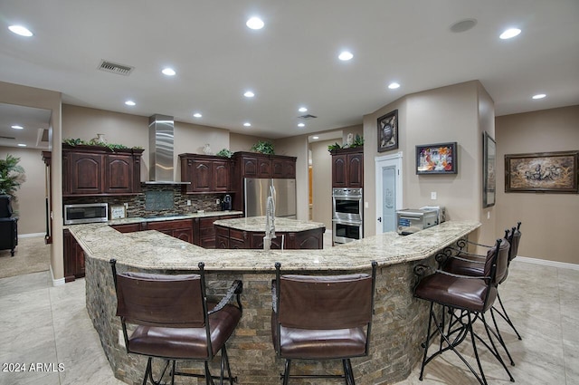 kitchen featuring appliances with stainless steel finishes, a breakfast bar, a large island, dark brown cabinetry, and wall chimney range hood