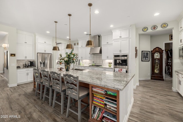 kitchen with appliances with stainless steel finishes, a large island with sink, white cabinets, and wall chimney range hood