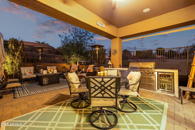 patio terrace at dusk featuring grilling area, an outdoor kitchen, beverage cooler, and a fire pit