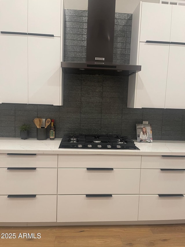 kitchen featuring white cabinets, decorative backsplash, light stone counters, black gas stovetop, and wall chimney range hood