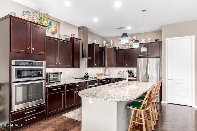 kitchen with stainless steel appliances, dark wood-type flooring, a sink, light stone countertops, and wall chimney exhaust hood