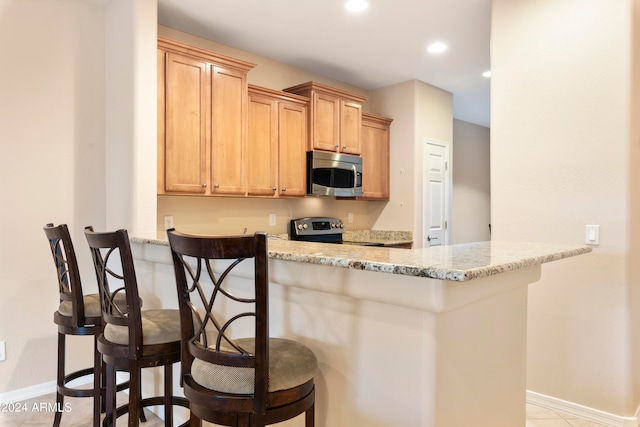 kitchen with light brown cabinetry, kitchen peninsula, stainless steel appliances, light stone counters, and a breakfast bar area