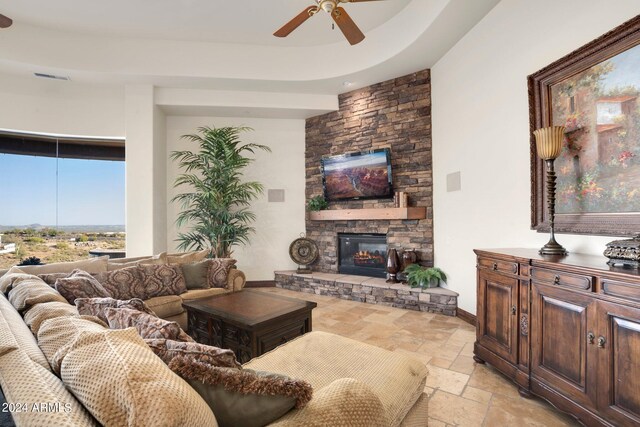 living area featuring ceiling fan, baseboards, a tray ceiling, a stone fireplace, and stone tile flooring