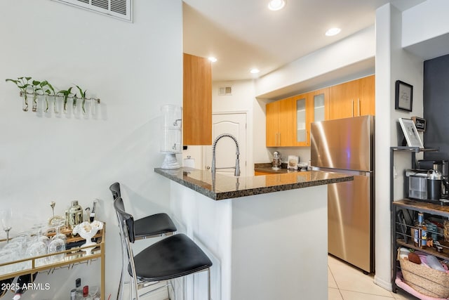 kitchen with kitchen peninsula, stainless steel fridge, light tile patterned flooring, dark stone countertops, and a breakfast bar area