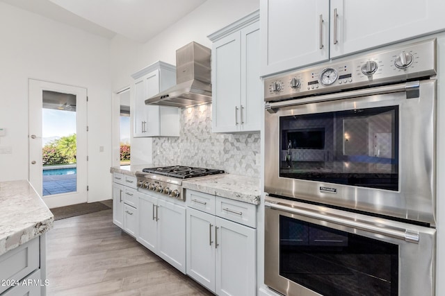 kitchen featuring light stone countertops, white cabinetry, wall chimney exhaust hood, stainless steel appliances, and backsplash