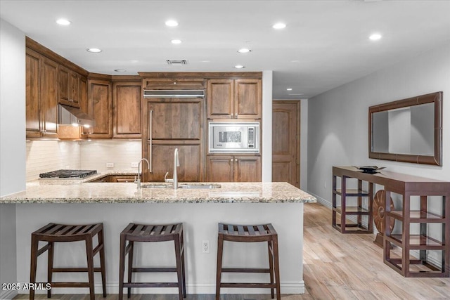 kitchen featuring sink, light stone counters, built in appliances, kitchen peninsula, and exhaust hood