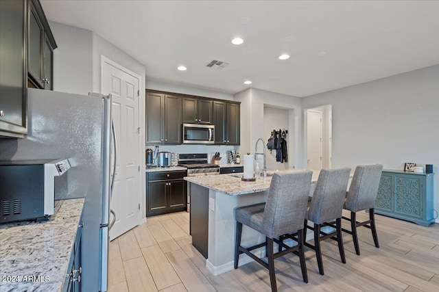 kitchen with light stone countertops, appliances with stainless steel finishes, light wood-type flooring, a kitchen breakfast bar, and a center island with sink